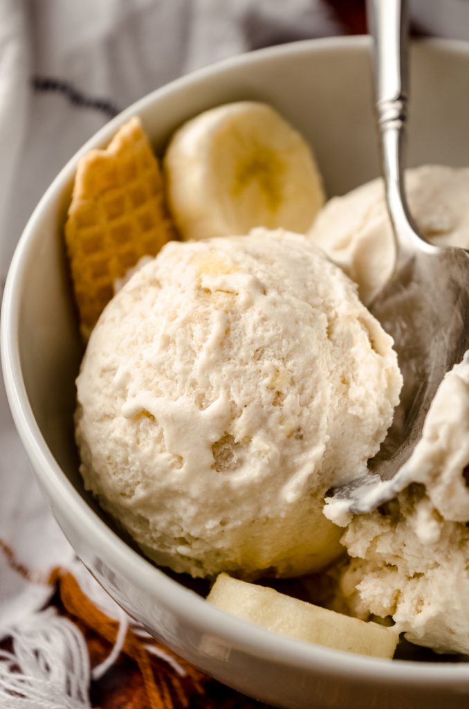 A bowl of roasted banana ice cream in a bowl with a spoon, banana slices, and pieces of waffle cone in it.