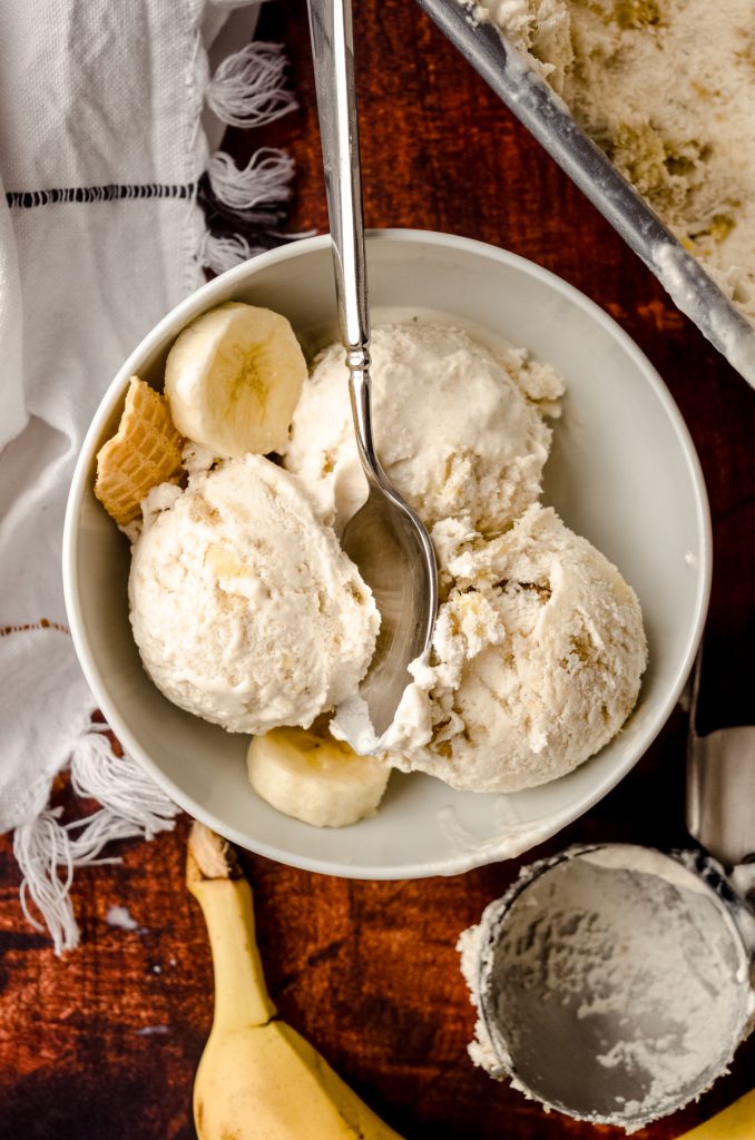 Aerial photo of a bowl of roasted banana ice cream in a bowl with a spoon, banana slices, and pieces of waffle cone in it.