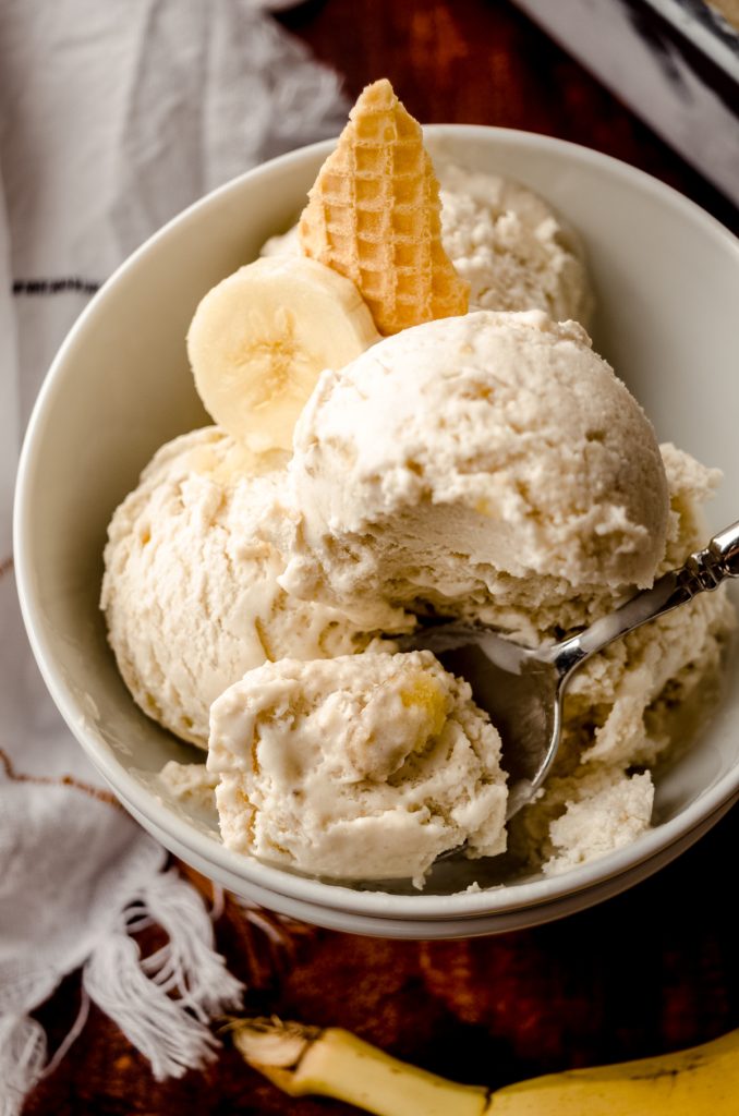 A bowl of roasted banana ice cream in a bowl with a spoon, banana slices, and pieces of waffle cone in it.