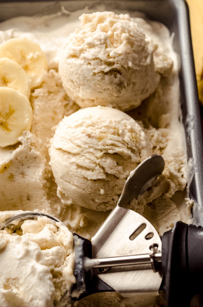 Roasted banana ice cream formed into a scoop sitting in a container of more ice cream with an ice cream scoop in the background.