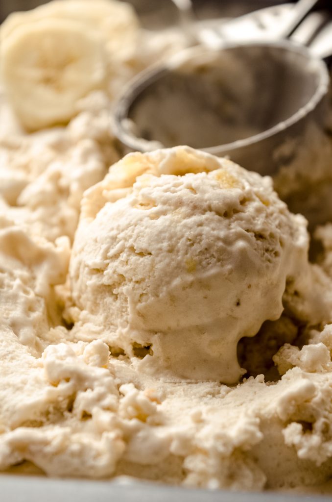 Roasted banana ice cream formed into a scoop sitting in a container of more ice cream with an ice cream scoop in the background.