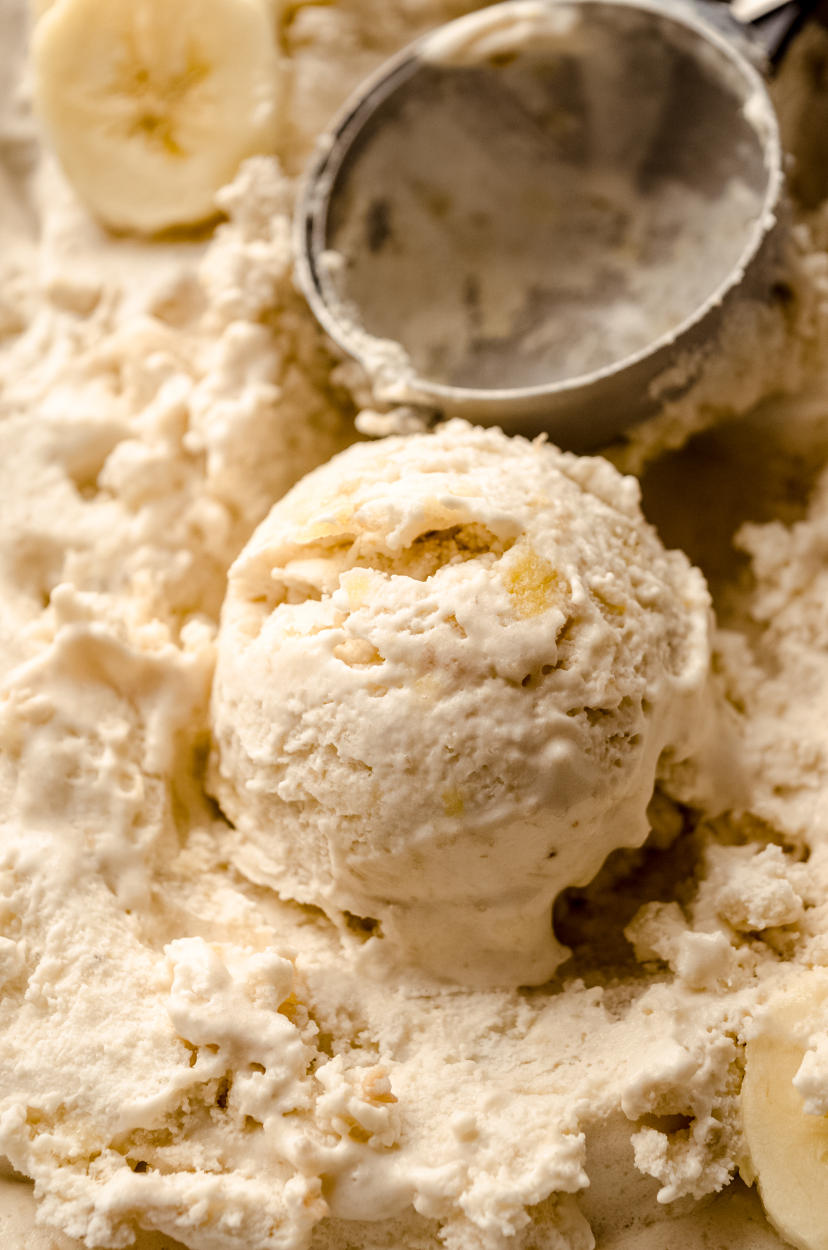 Roasted banana ice cream formed into a scoop sitting in a container of more ice cream with an ice cream scoop in the background.