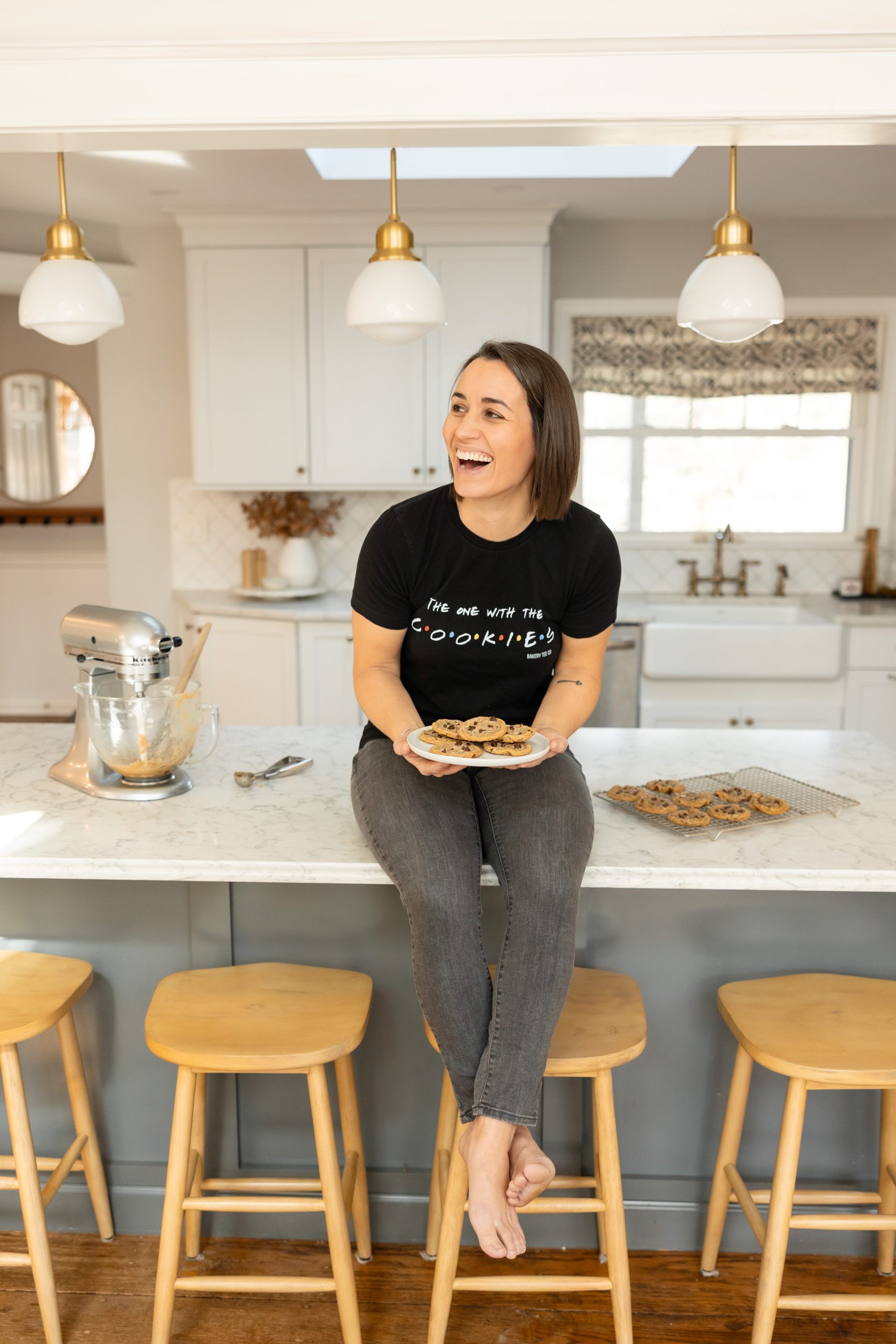 A woman sitting on a kitchen counter holding a plate of cookies.