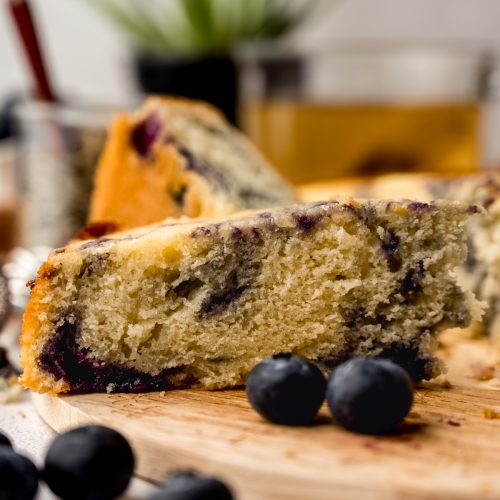 A blueberry tea cake sitting on a wooden trivet with blueberries in the foreground and a plant and loose leaf tea in the background.