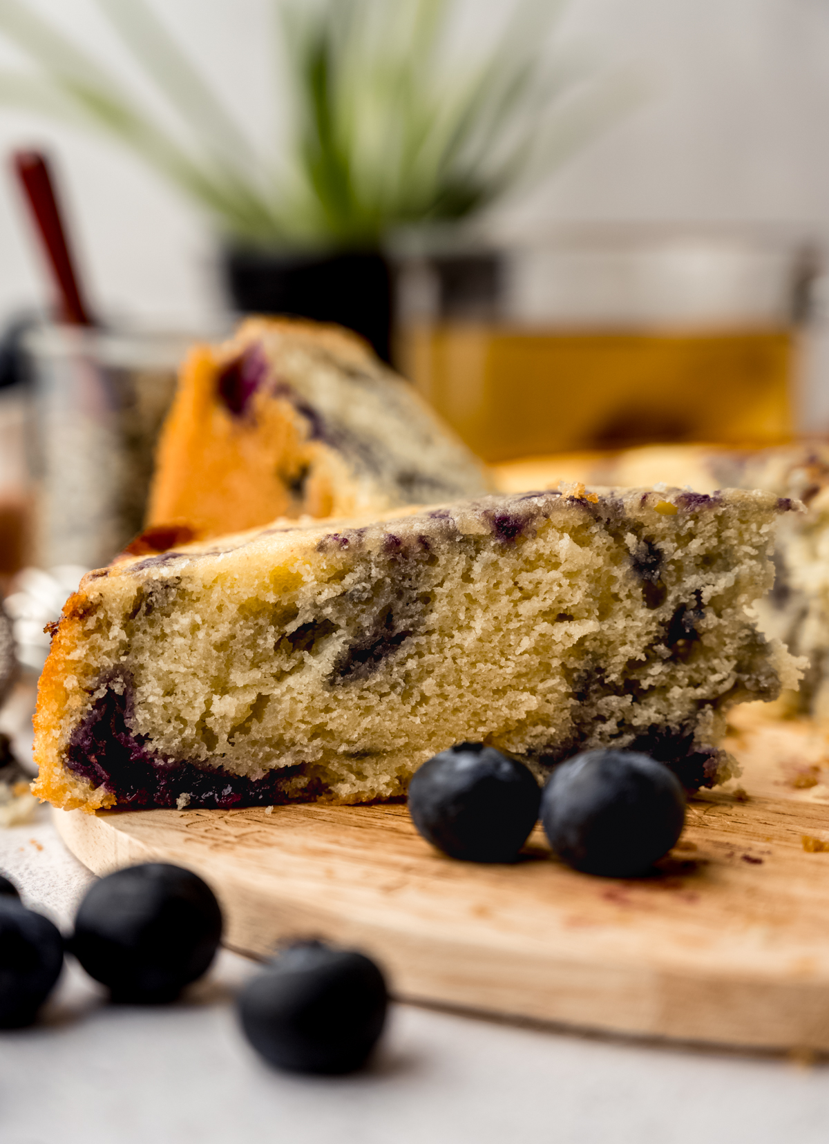 A blueberry tea cake sitting on a wooden trivet with blueberries in the foreground and a plant and loose leaf tea in the background.