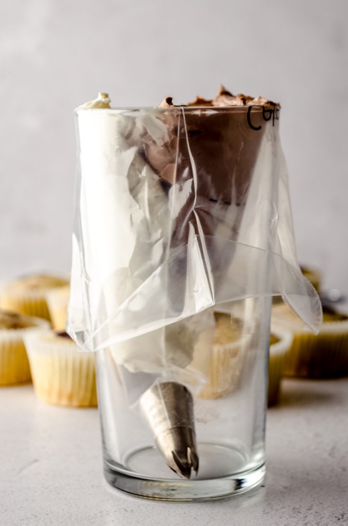 A piping bag fitted with a piping tip and sitting in a tall glass with white and brown frosting in it. 