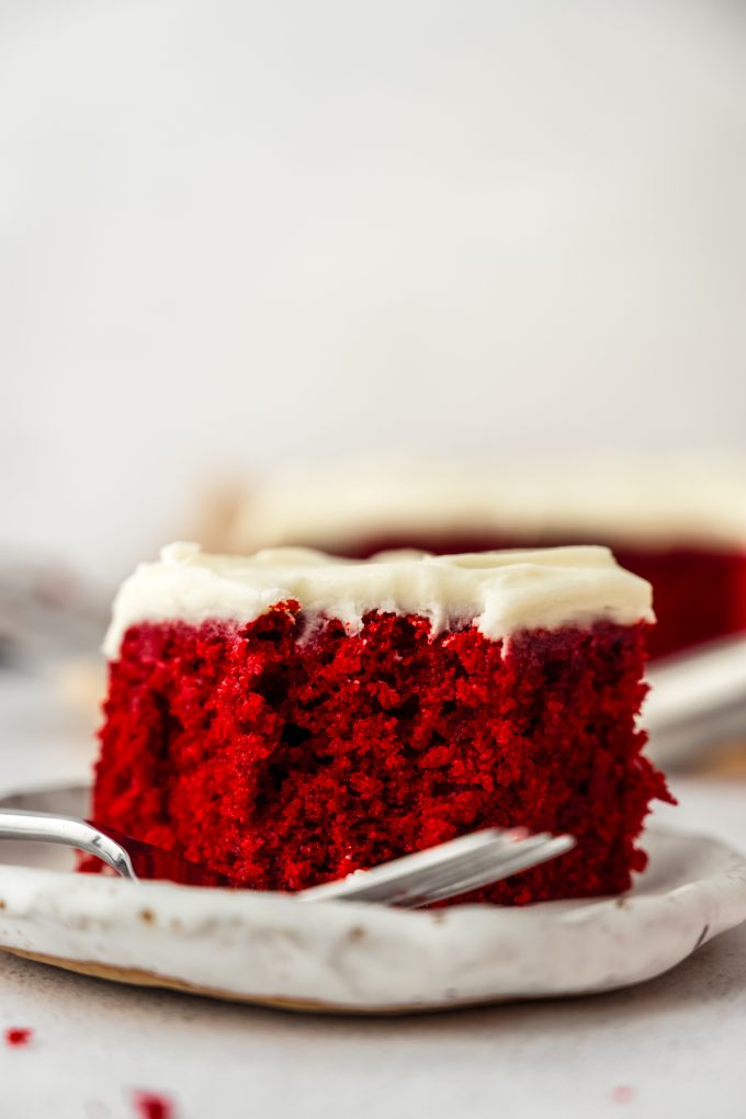 A slice of red velvet sheet cake on a plate with a fork and a bite has been taken out of it.