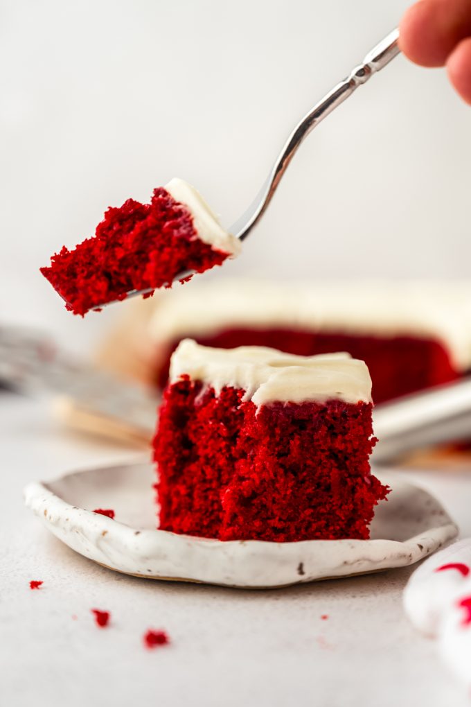 Someone is removing a bite from a slice of red velvet sheet cake on a plate with a fork.