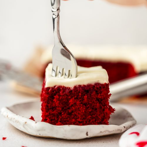 A slice of red velvet sheet cake on a plate with someone's hand using a fork to take a bite.