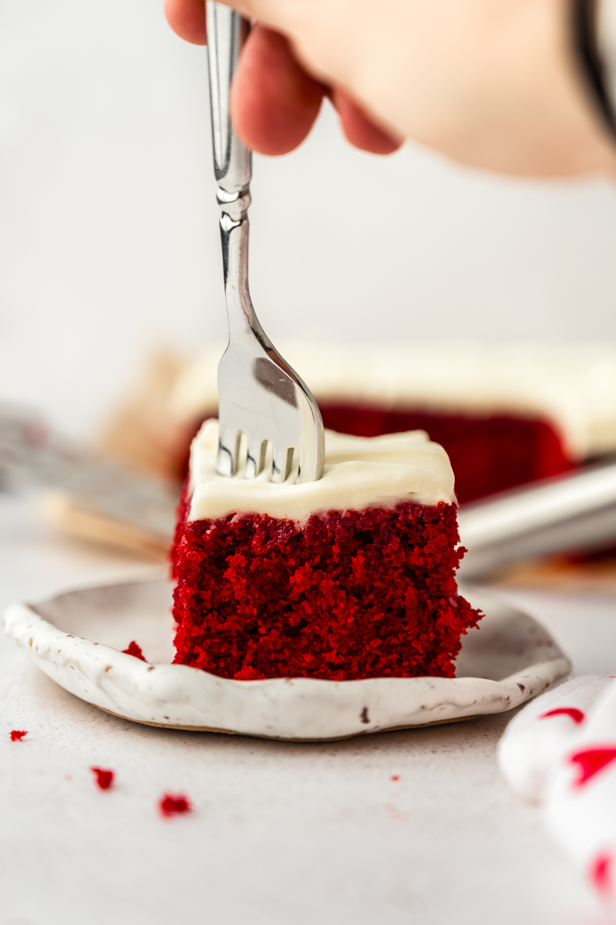 A slice of red velvet sheet cake on a plate with someone's hand using a fork to take a bite.