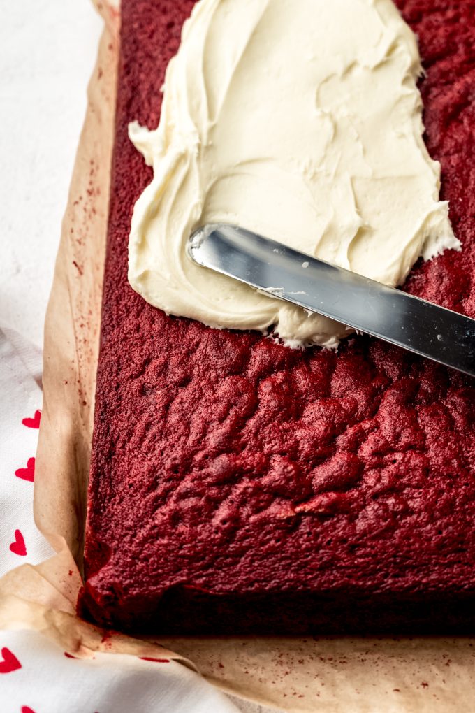 Someone is using a large offset metal spatula to spread cream cheese frosting onto a red velvet sheet cake.