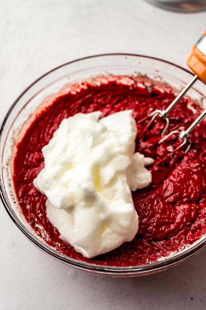 A bowl of red velvet cake batter in a bowl with electric beaters and a heaping pile of whipped egg whites sits on top ready to be folded into the batter.