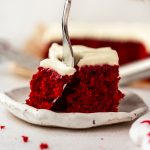 A slice of red velvet sheet cake on a plate with someone's hand using a fork to take a bite.