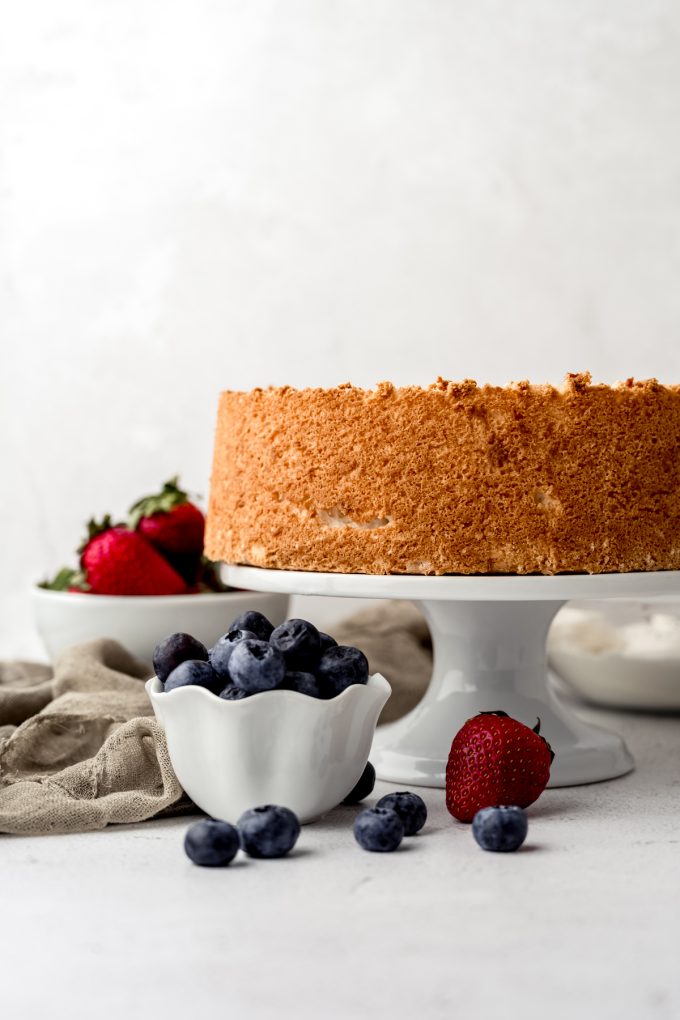 An angel food cake on a cake stand with blueberries and strawberries around it.