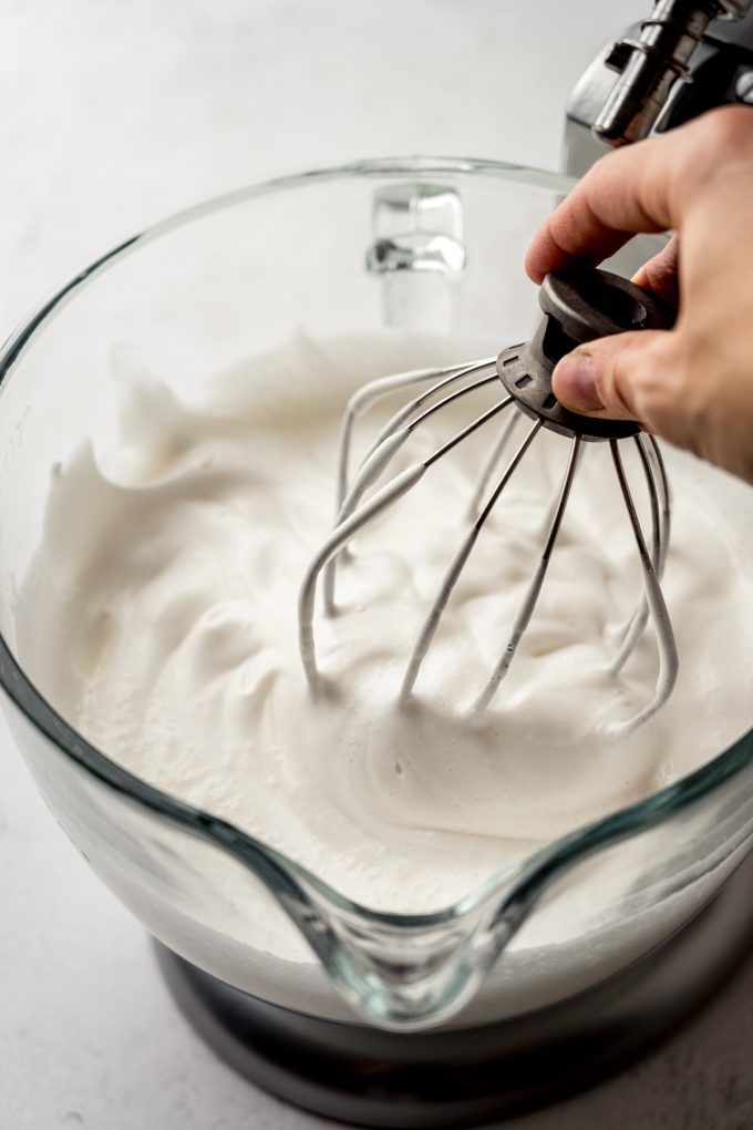 Someone is holding the whisk attachment of a stand mixer in the bowl with angel food cake batter in it.