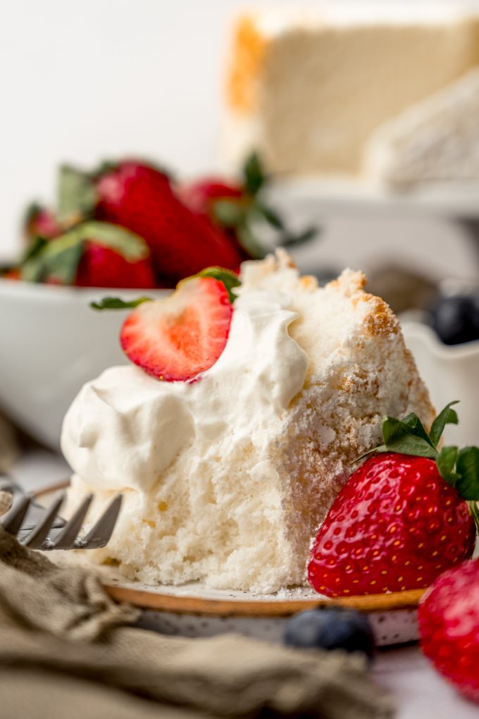 A slice of angel food cake with whipped cream and strawberries on it sitting on a plate with a fork and a bite taken out of it.