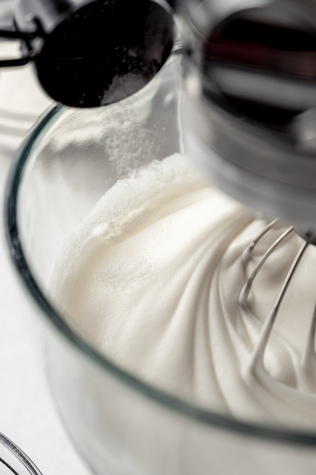 Someone is pouring sugar into the bowl of a stand mixer that has meringue in it.