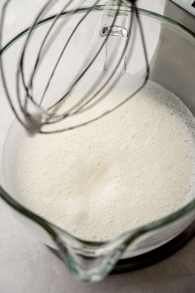 The bowl of a stand mixer fitted with a whisk attachment with whipped egg whites in the bowl that are foamy.