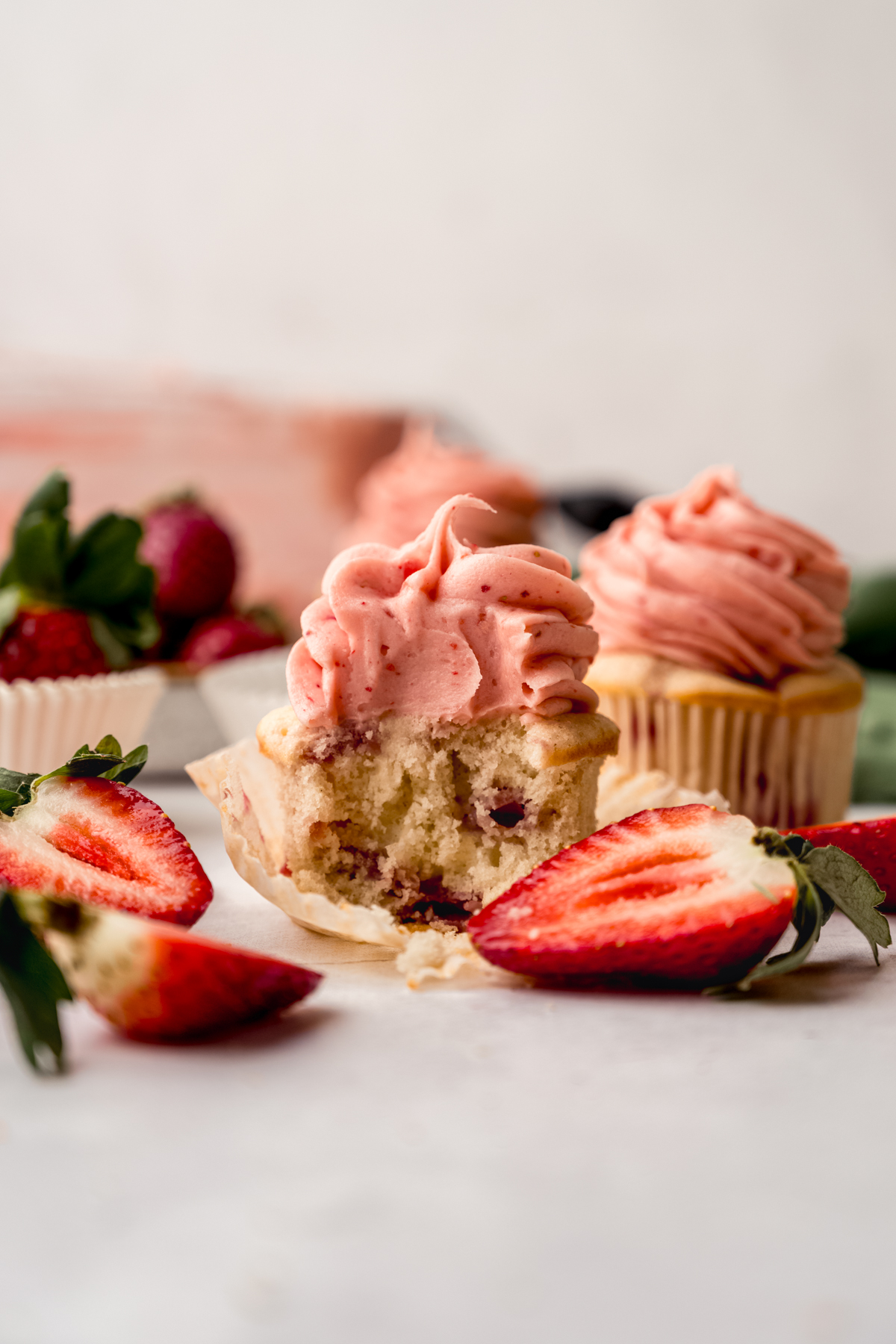 A frosted strawberry cupcake with a bite taken out of it sitting on a surface.