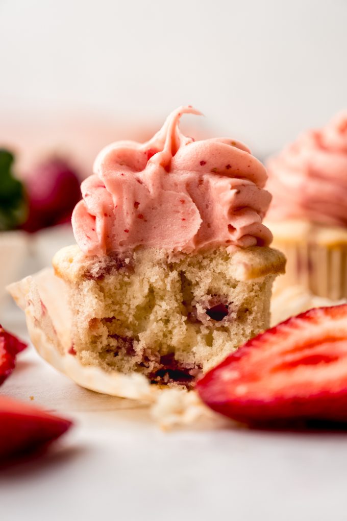 A frosted strawberry cupcake with a bite taken out of it sitting on a surface.