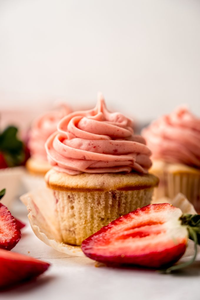 A frosted strawberry cupcake with the wrapper pulled down sitting on a surface.