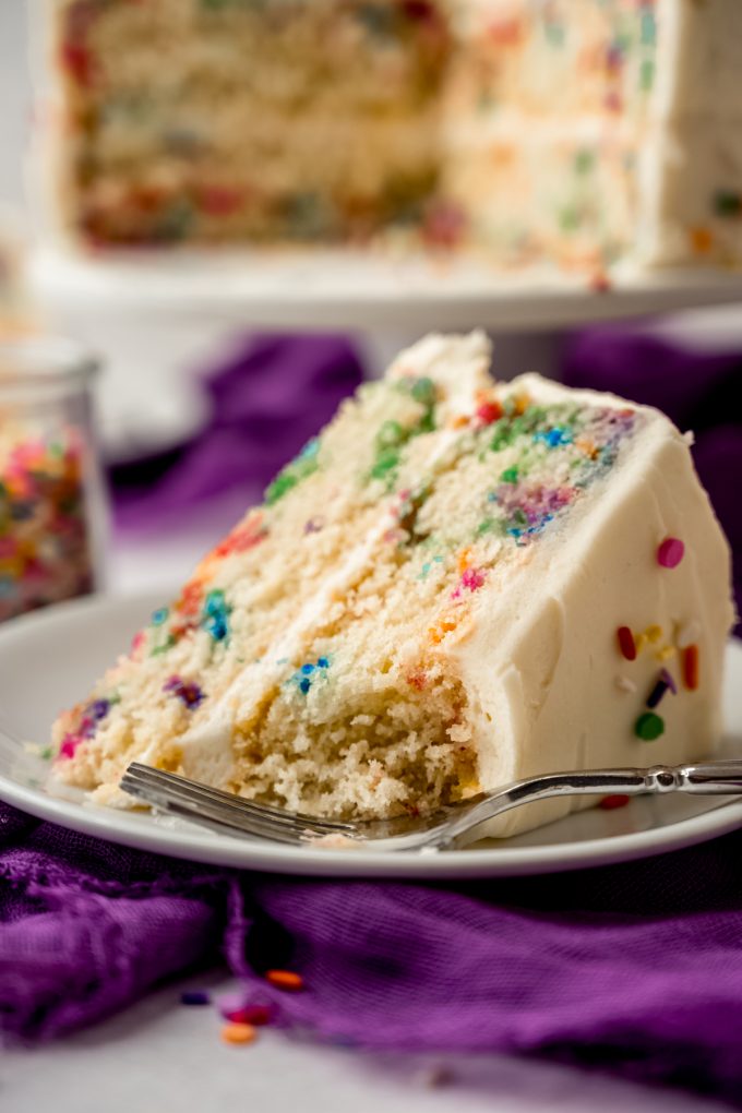 A slice of confetti layer cake on a plate with a fork and a bite taken out of the cake.