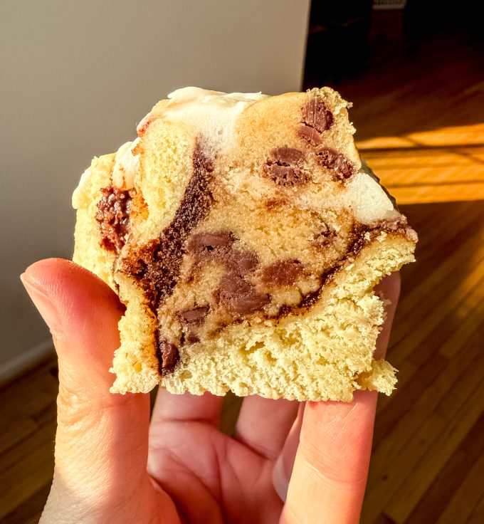Someone is holding a cookie dough cinnamon roll in their hand and the roll has been sliced in half and allowed to cool so you can see all of the cookie dough in the center.