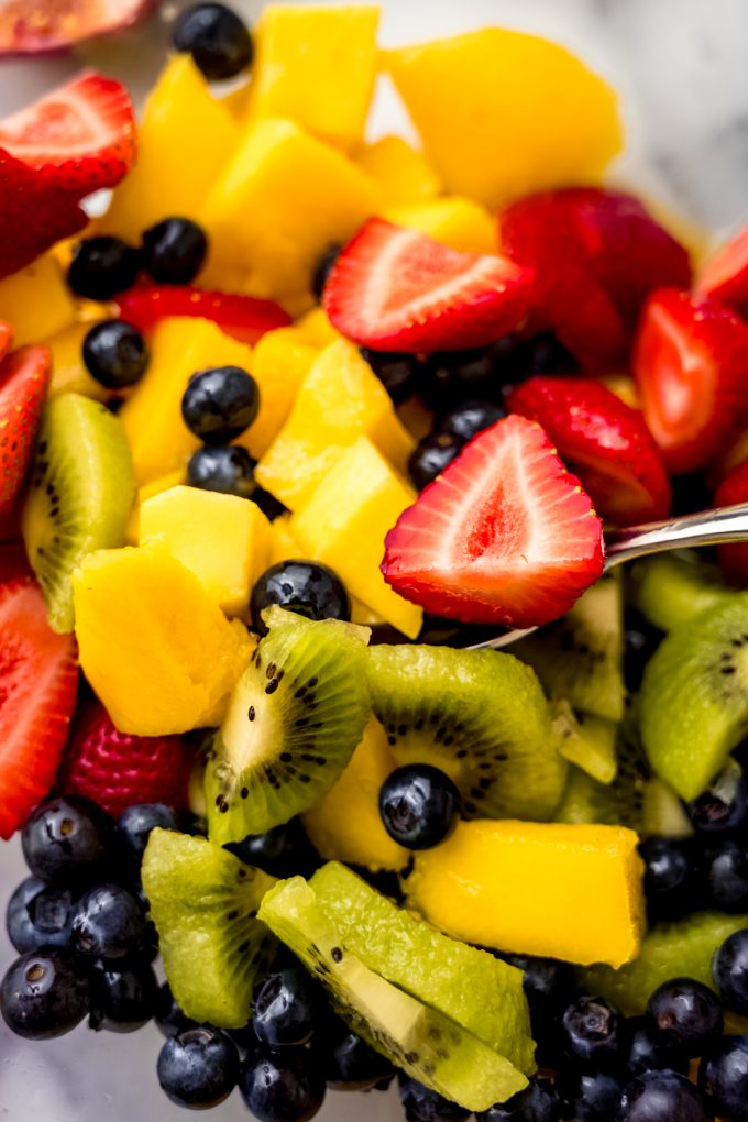 Someone is using a spoon to stir a large bowl of cut fruit to make fruit salad.