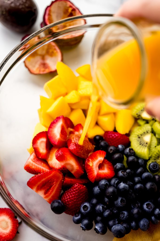 Someone is pouring passion fruit dressing onto cut fruit in a large glass bowl.