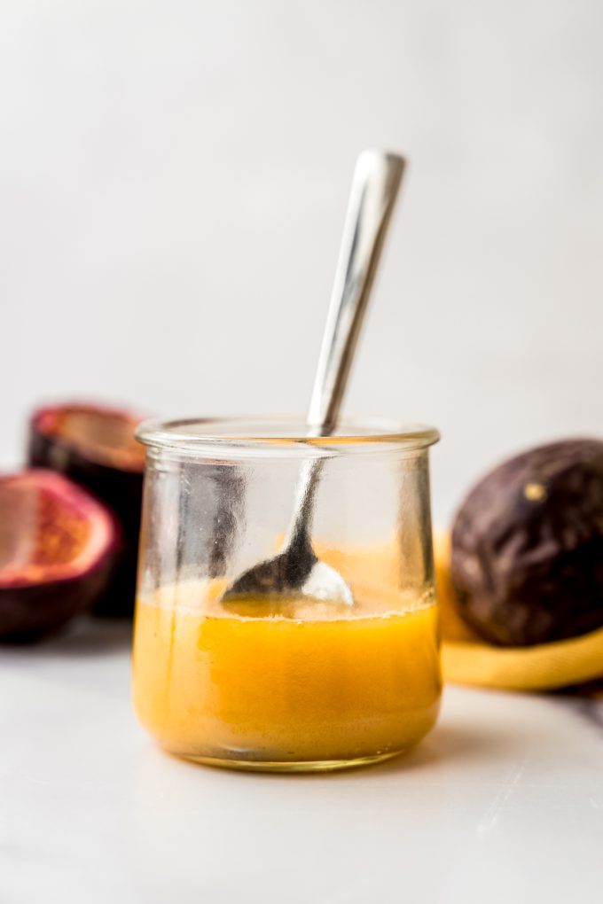 Passion fruit dressing in a small glass container with a spoon and passion fruits in the background.