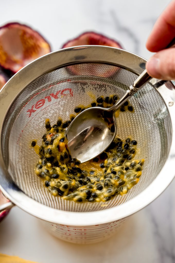 Someone is pressing passion fruit pulp through a mesh sieve over a glass measuring cup with a spoon.