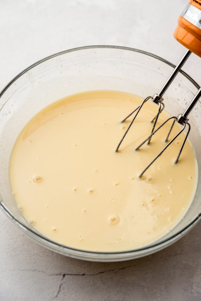 Pudding mixture for banana pudding in a large glass bowl with electric hand mixer beaters in it.