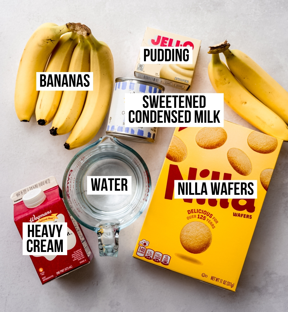 Aerial photo of ingredients to make banana pudding on a surface and the photo has text overlay labeling each ingredient.