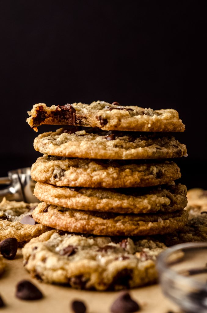 A stack of brown butter chocolate chip cookies on parchment paper with chocolate chips in the foreground and a bite taken out of the cookie on the top of the stack.