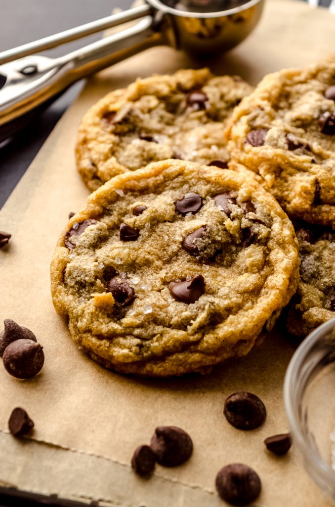 Brown butter chocolate chip cookies on a piece of parchment paper with a cookie scoop in the background and chocolate chips in the foreground.