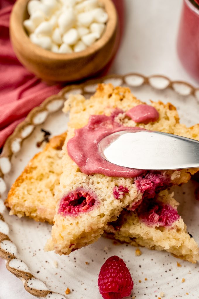 Someone is using a spreading knife to put raspberry curd onto a raspberry white chocolate chip scone.