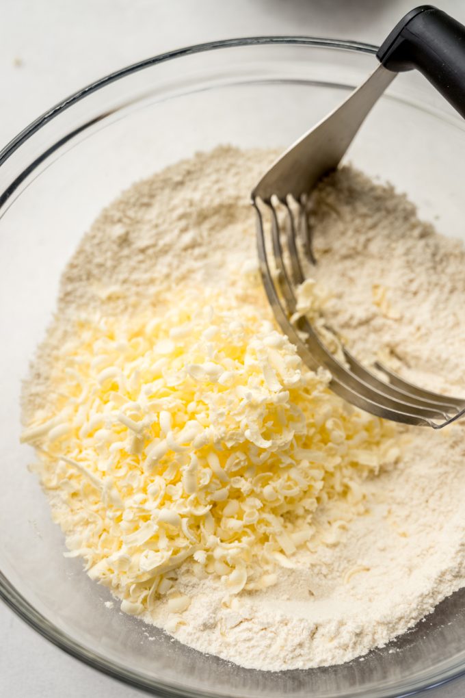 The dough for scones in a bowl with grated butter and a pastry cutter in it.