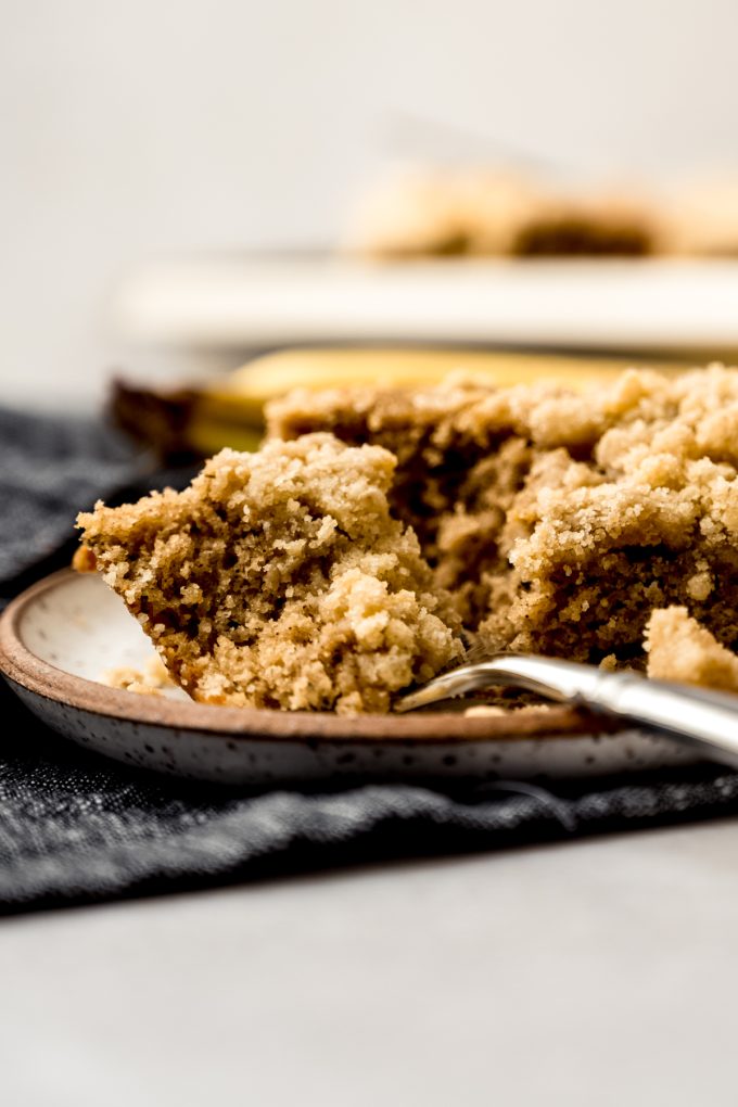 A slice of banana crumb cake on a plate with a fork and a bite taken out of the cake.
