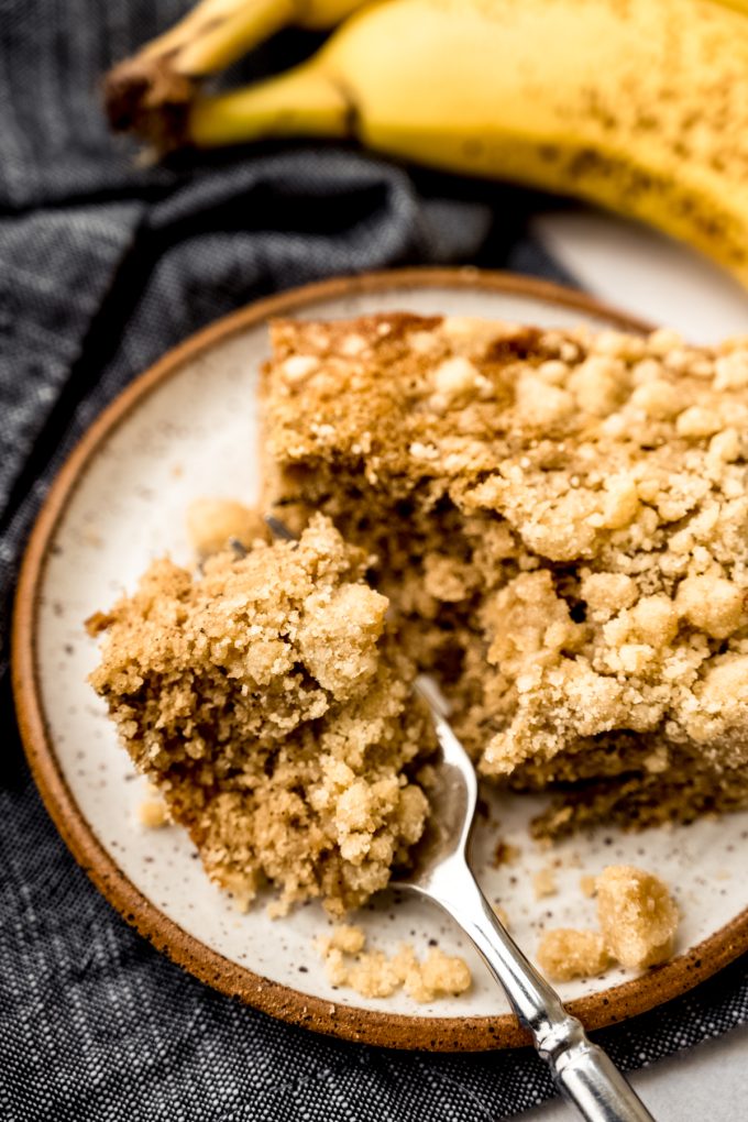 A slice of banana crumb cake on a plate with a fork and a bite taken out of the cake.
