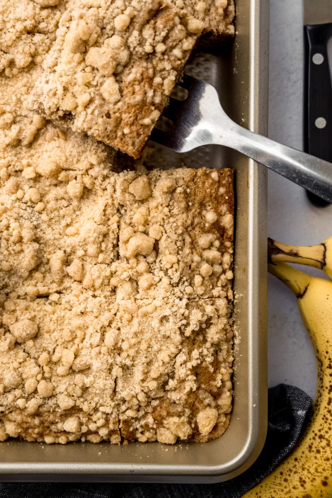 Aerial photo of sliced banana crumb cake in a pan with a spatula.