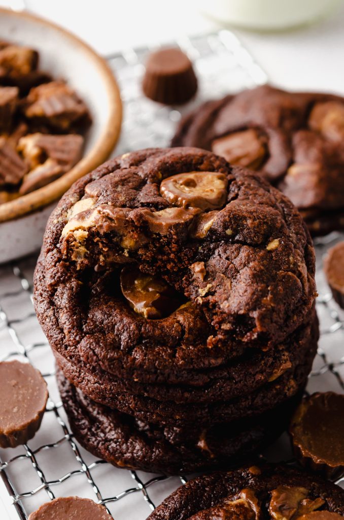 A stack of chocolate peanut butter cookies on a cooling rack with peanut butter cups around it and you can see a jug of milk in the background.