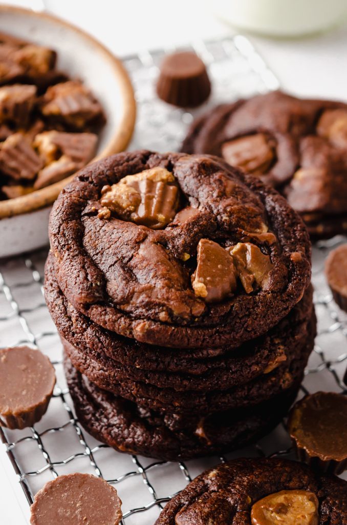 A stack of chocolate peanut butter cookies on a cooling rack with peanut butter cups around it and you can see a jug of milk in the background.