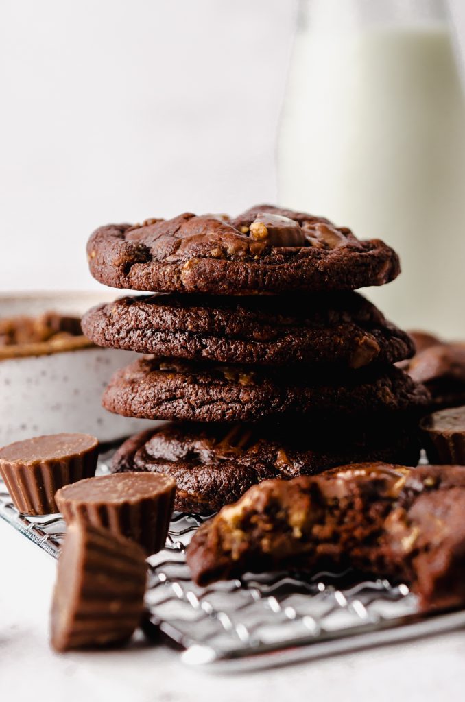 A stack of chocolate peanut butter cookies on a cooling rack with peanut butter cups around it and you can see a jug of milk in the background.