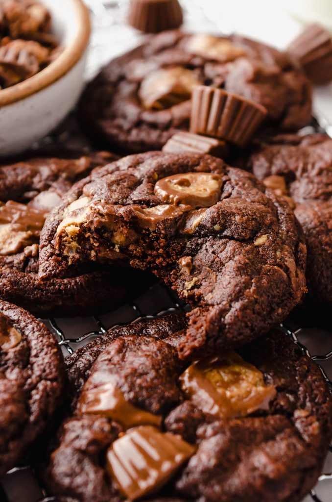 A photo of a chocolate peanut butter cup cookie with a bite taken out of it on a surface with peanut butter cups around it.