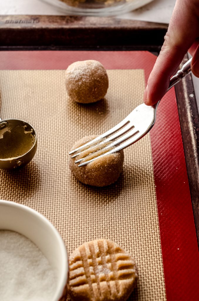 Someone is using a fork to make a criss-cross pattern on a peanut butter cookie dough ball.