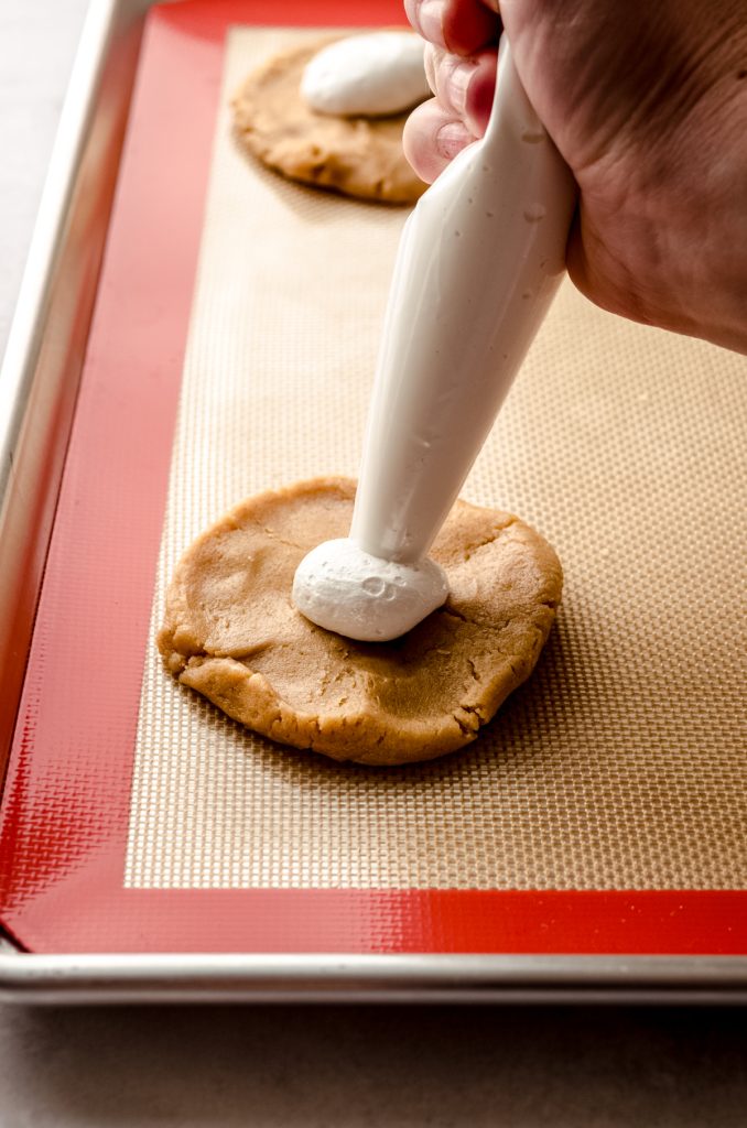 Someone is piping marshmallow fluff onto a disc of peanut butter cookie dough to make fluffernutter cookies.