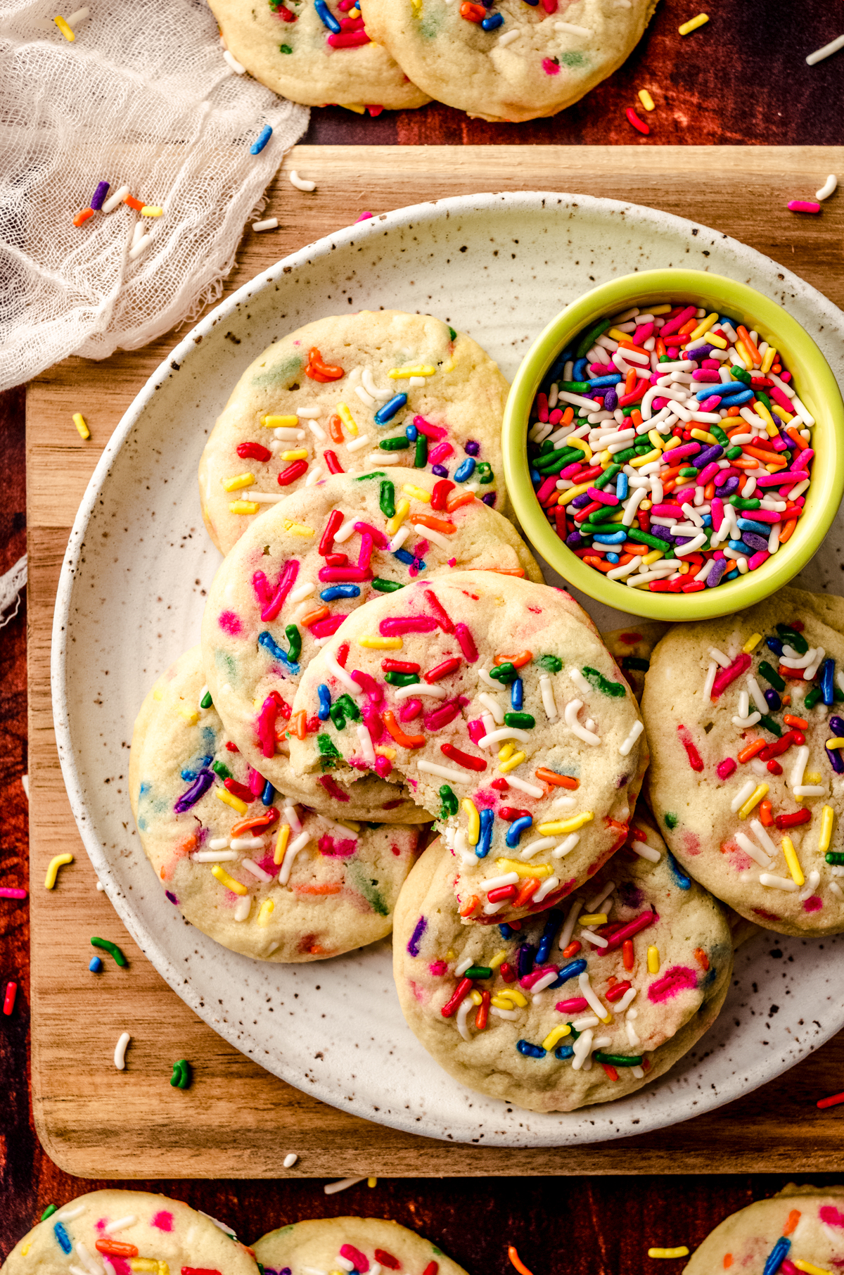 Aerial photo of sprinkle sugar cookies on a plate with a bowl of sprinkles and sprinkles scattered all around it and a bite has been taken out of one of the cookies.