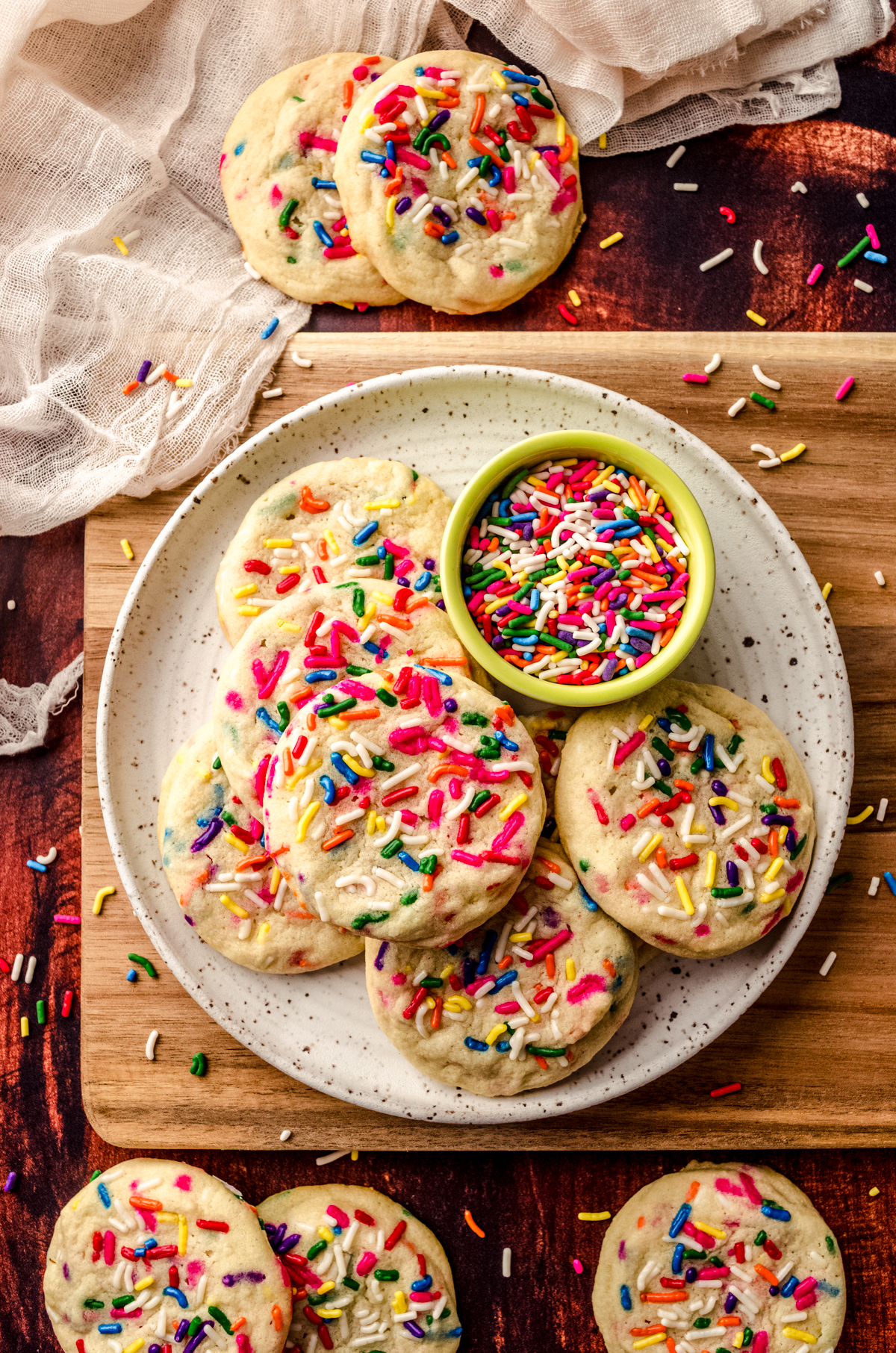 Aerial photo of sprinkle sugar cookies on a plate with a bowl of sprinkles and sprinkles scattered all around it.