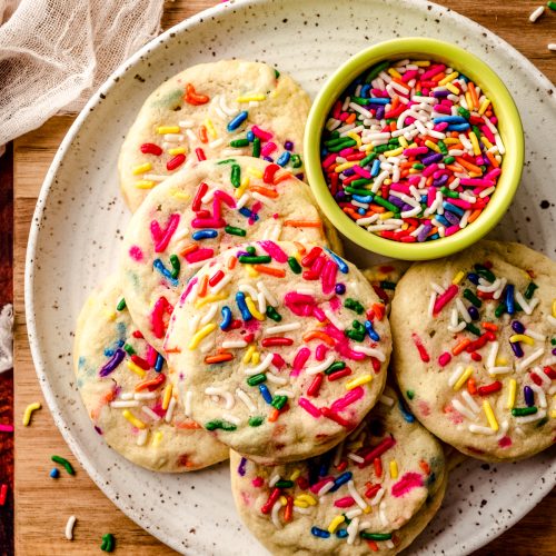 Aerial photo of sprinkle sugar cookies on a plate with a bowl of sprinkles and sprinkles scattered all around it.