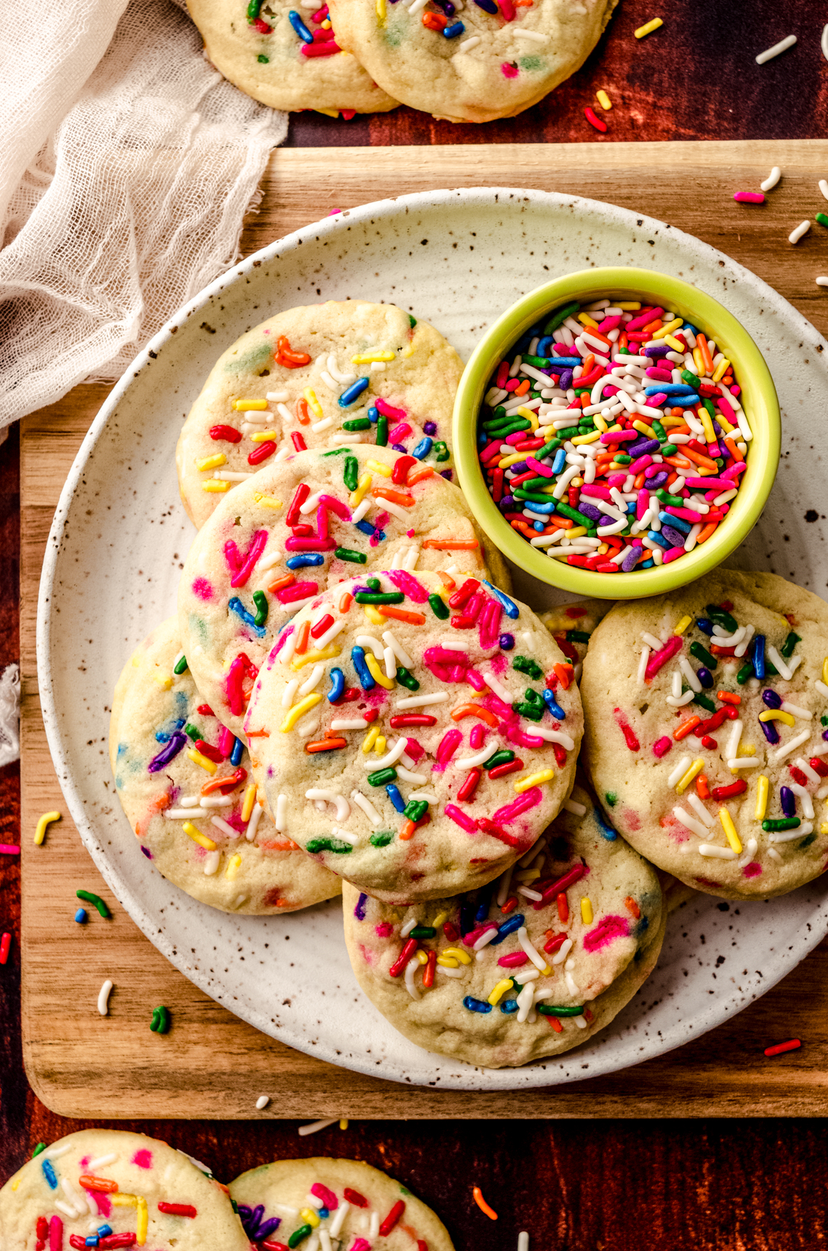 Aerial photo of sprinkle sugar cookies on a plate with a bowl of sprinkles and sprinkles scattered all around it.